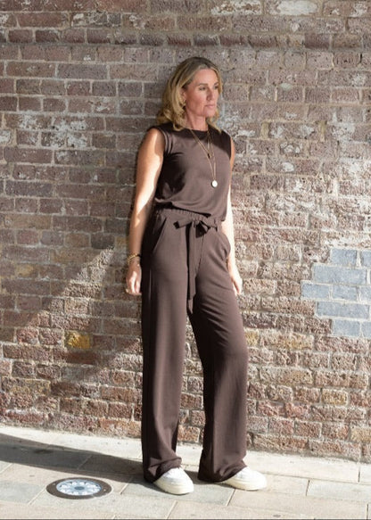 Woman in a brown jumpsuit standing against a brick wall.